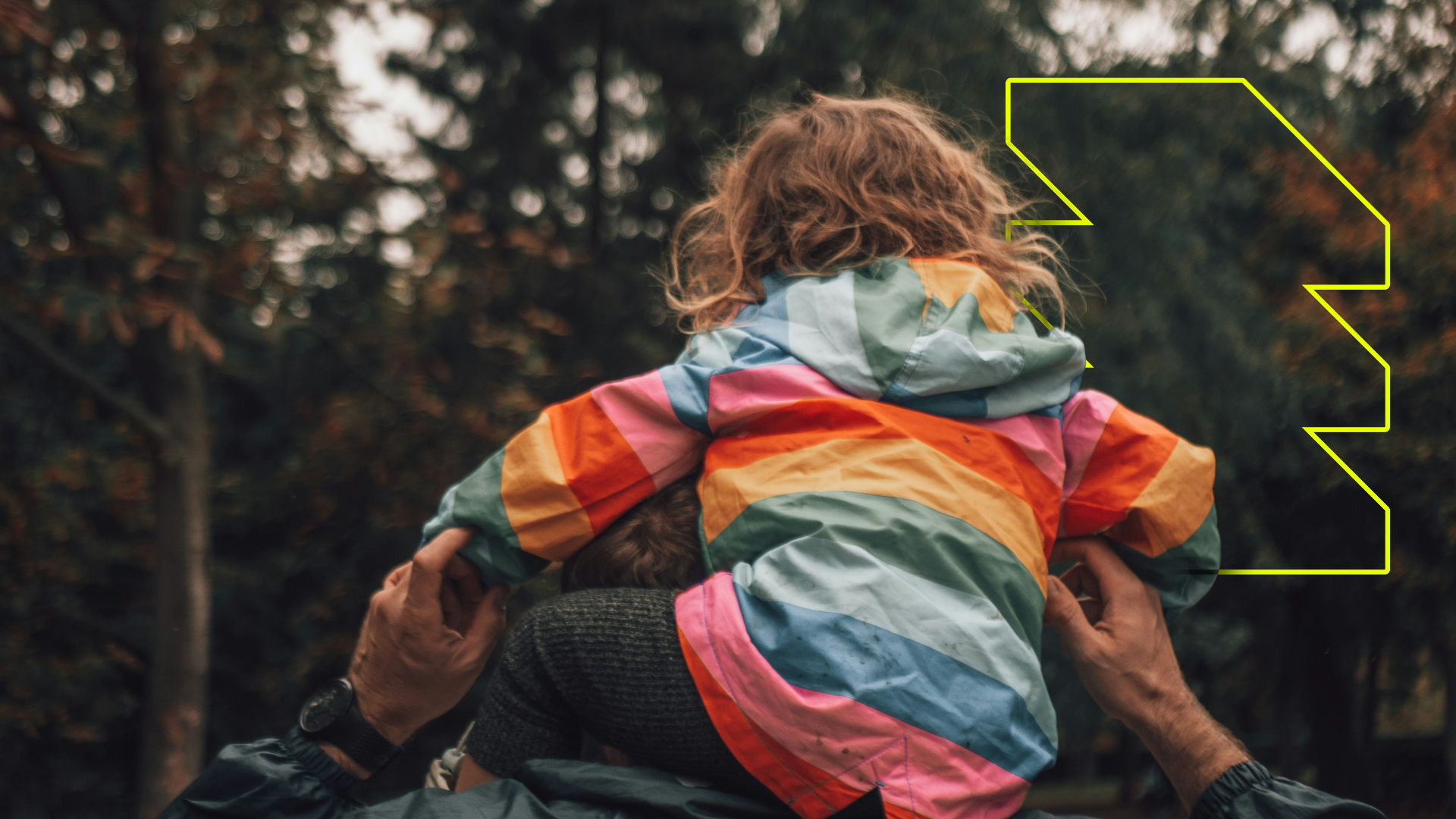 A child in a colourful striped waterproof jacket on an adult's shoulders, walking outdoors among trees